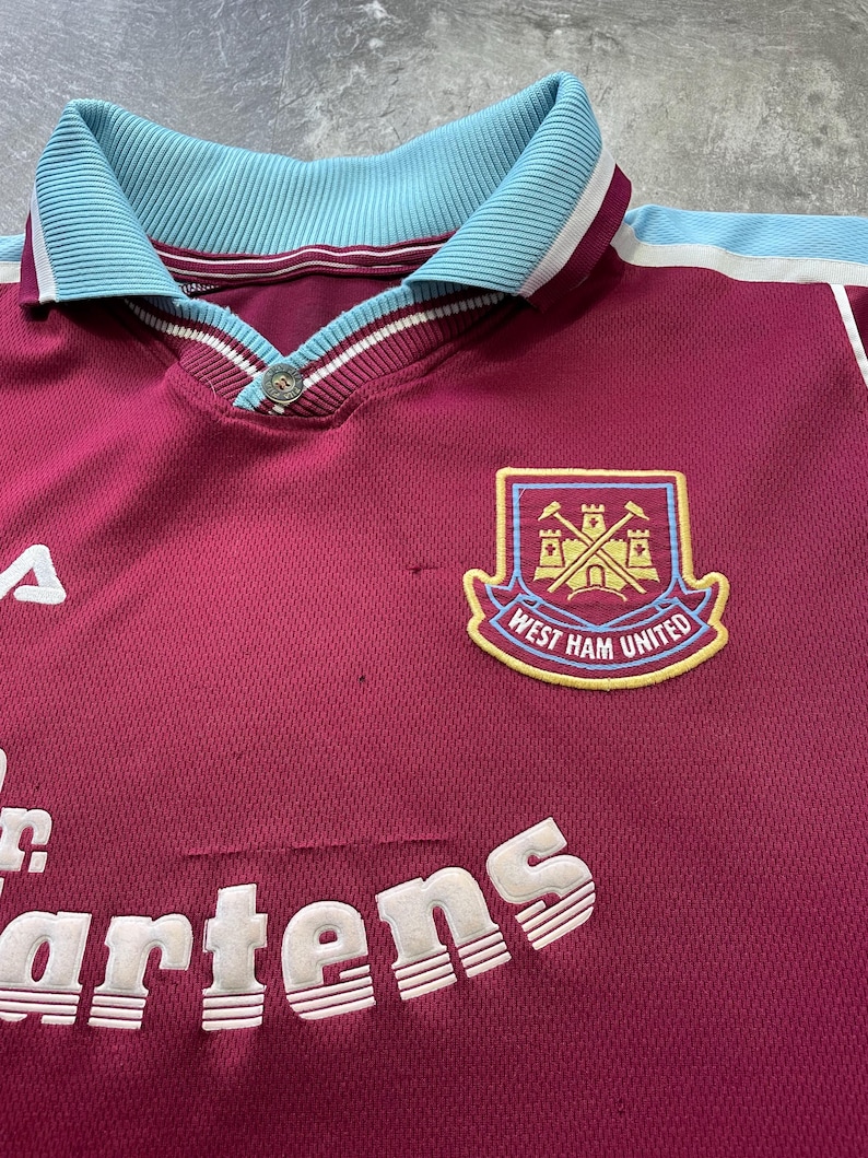 May include: Maroon and light blue West Ham United football shirt. Features a collared neckline, button closure, and the club crest. The Dr. Martens logo is also visible.