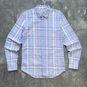 May include: A long-sleeve button-down shirt featuring a light blue and white plaid pattern. The shirt has a standard collar, a front button placket, and barrel cuffs. An Acne Studios brand label is visible on the inside of the collar neckline.