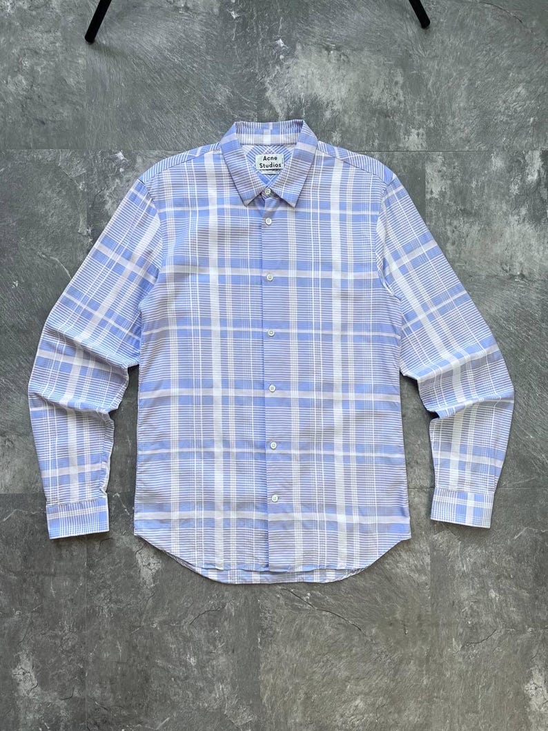 May include: A long-sleeve button-down shirt featuring a light blue and white plaid pattern. The shirt has a standard collar, a front button placket, and barrel cuffs. An Acne Studios brand label is visible on the inside of the collar neckline.