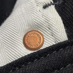 May include: Close-up of a copper button with the text "Acne Studios" embossed. The button is on black fabric with a white herringbone lining.