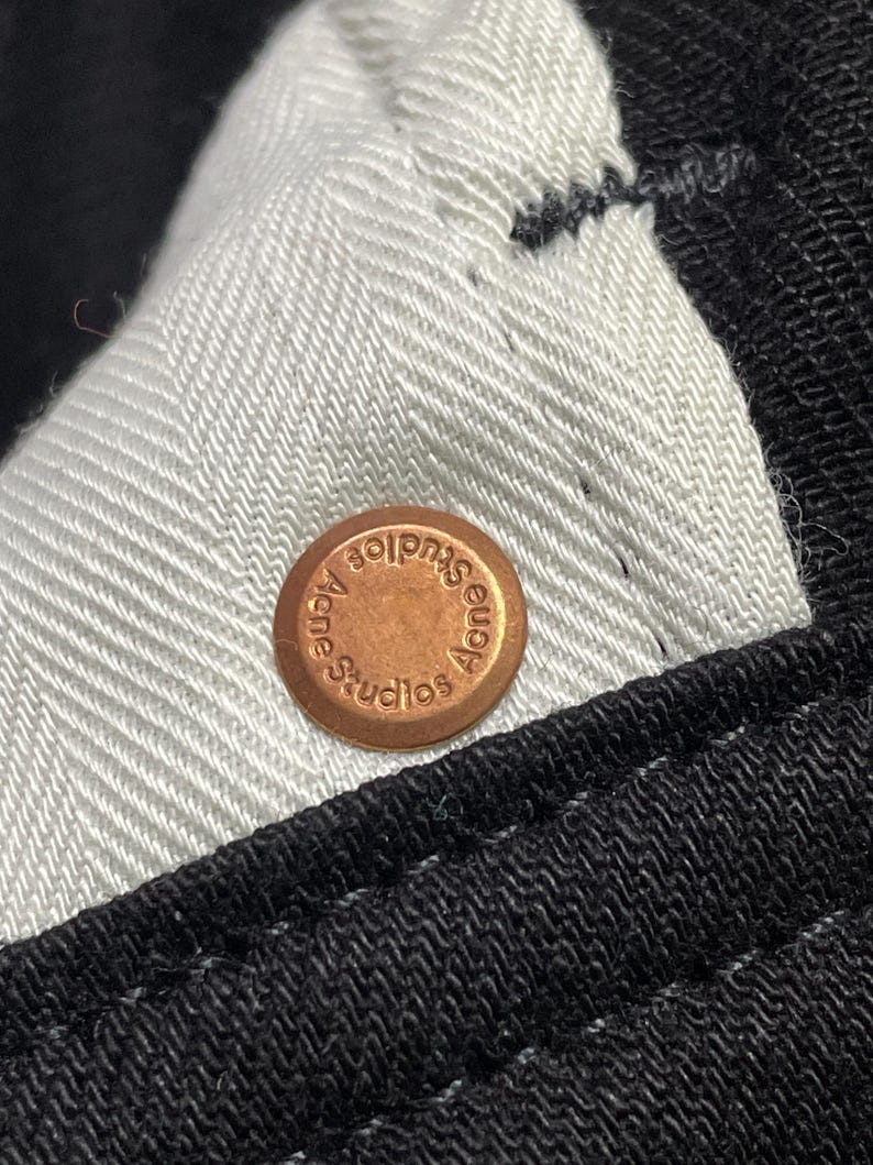 May include: Close-up of a copper button with the text "Acne Studios" embossed. The button is on black fabric with a white herringbone lining.