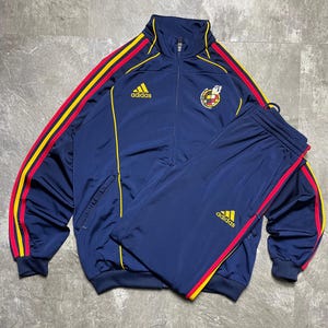 May include: Navy blue Adidas track suit with yellow and red stripes. The jacket has a full zipper and the Adidas logo on the chest. The matching pants also have the Adidas logo. The track suit has a Spanish national team emblem.
