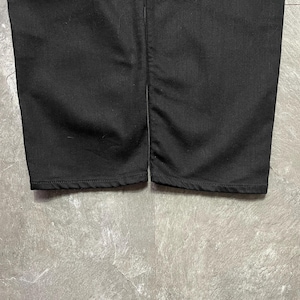 May include: Close-up of two black trouser legs. The fabric appears to be a sturdy material, with visible seams at the bottom hems. The trousers are set against a grey, textured surface.