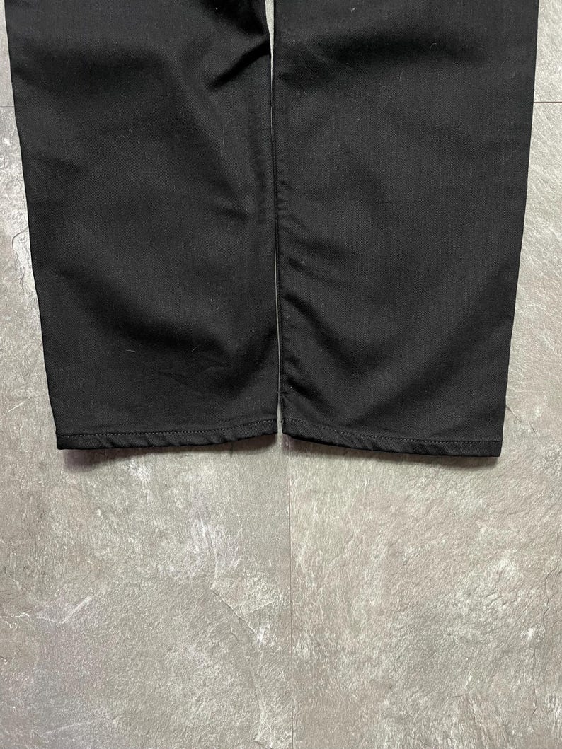 May include: Close-up of two black trouser legs. The fabric appears to be a sturdy material, with visible seams at the bottom hems. The trousers are set against a grey, textured surface.
