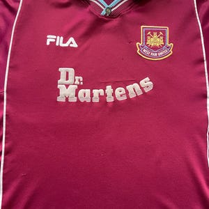 May include: Maroon and light blue football shirt with the Fila logo and Dr. Martens text. The shirt features a West Ham United crest on the chest. The collar and shoulder accents are light blue.