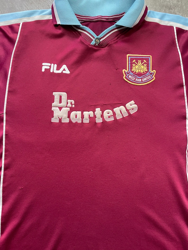 May include: Maroon and light blue football shirt with the Fila logo and Dr. Martens text. The shirt features a West Ham United crest on the chest. The collar and shoulder accents are light blue.