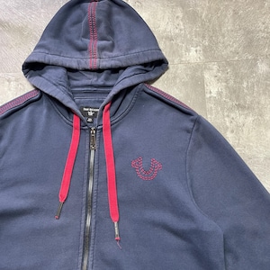 May include: Navy blue zip-up hoodie with a red drawstring and accents. Features a stitched red design on the hood and shoulders, and a stitched logo on the chest. The hoodie has a full zipper and a front pocket.