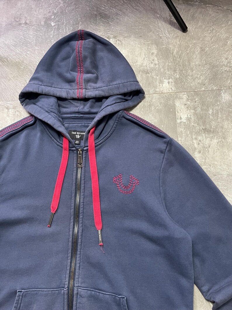 May include: Navy blue zip-up hoodie with a red drawstring and accents. Features a stitched red design on the hood and shoulders, and a stitched logo on the chest. The hoodie has a full zipper and a front pocket.