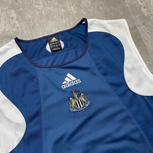 K&ouml;nnte beinhalten: Ein marineblaues Adidas-Tanktop-Trikot mit wei&szlig;en Akzenten. Das Trikot zeigt das Adidas-Logo und das Wappen von Newcastle United. Der Stoff hat eine strukturierte Optik, und der Halsausschnitt ist mit einem dunkelblauen Rand versehen.