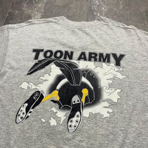 Puede incluir: Camiseta gris con el texto "TOON ARMY" en letras negras. La camiseta presenta un dibujo animado de un p&aacute;jaro negro con patas amarillas y botas de f&uacute;tbol. El p&aacute;jaro parece estar rompiendo un fondo blanco y gris.