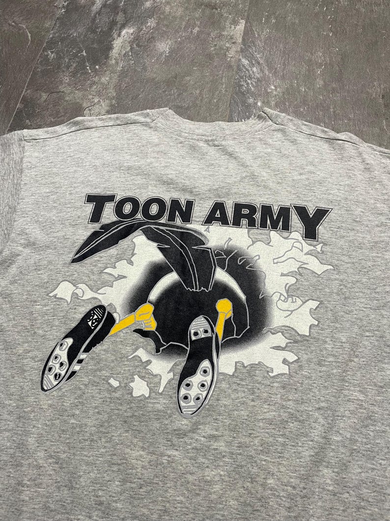 Puede incluir: Camiseta gris con el texto "TOON ARMY" en letras negras. La camiseta presenta un dibujo animado de un p&aacute;jaro negro con patas amarillas y botas de f&uacute;tbol. El p&aacute;jaro parece estar rompiendo un fondo blanco y gris.