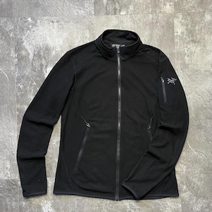 May include: Black full-zip jacket with a stand-up collar. The jacket features two side pockets and a small pocket on the upper left sleeve. A brand logo is visible on the upper left sleeve. The jacket is made of a textured fabric.