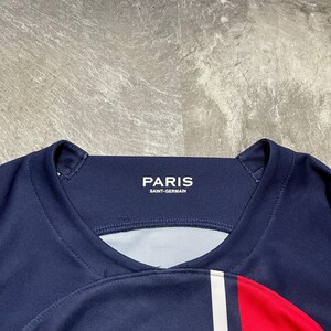 May include: A dark blue athletic jersey featuring a ribbed crew neckline with the white text PARIS SAINT-GERMAIN printed on the inner collar. The garment includes a vertical white stripe and a red panel on the right side of the chest area.