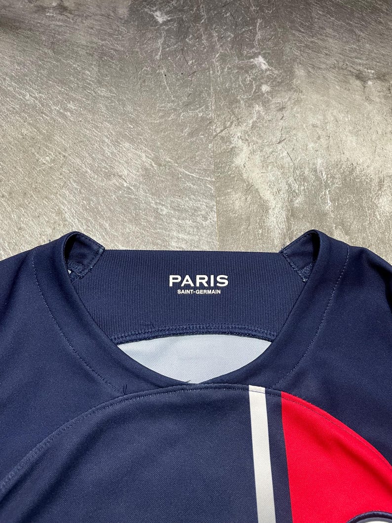 May include: A dark blue athletic jersey featuring a ribbed crew neckline with the white text PARIS SAINT-GERMAIN printed on the inner collar. The garment includes a vertical white stripe and a red panel on the right side of the chest area.
