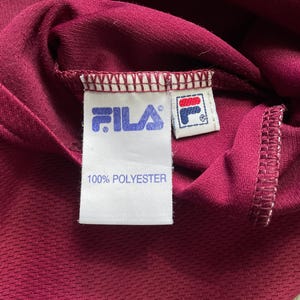 May include: Close-up of a burgundy athletic shirt with the Fila logo and a tag that reads "100% POLYESTER". The fabric has a textured appearance.