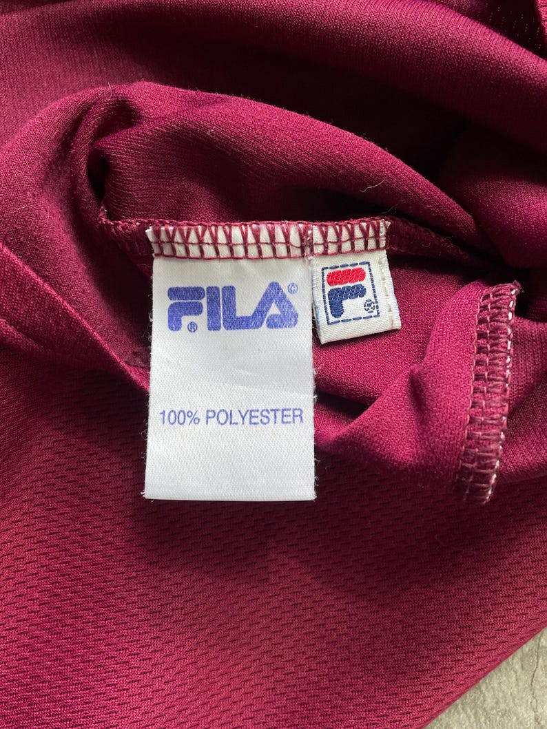 May include: Close-up of a burgundy athletic shirt with the Fila logo and a tag that reads "100% POLYESTER". The fabric has a textured appearance.
