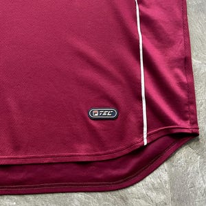 May include: Maroon athletic shirt with a white stripe down the side. The shirt has a mesh texture and a small logo near the bottom. The fabric appears to be a lightweight, breathable material, suitable for sports or casual wear.