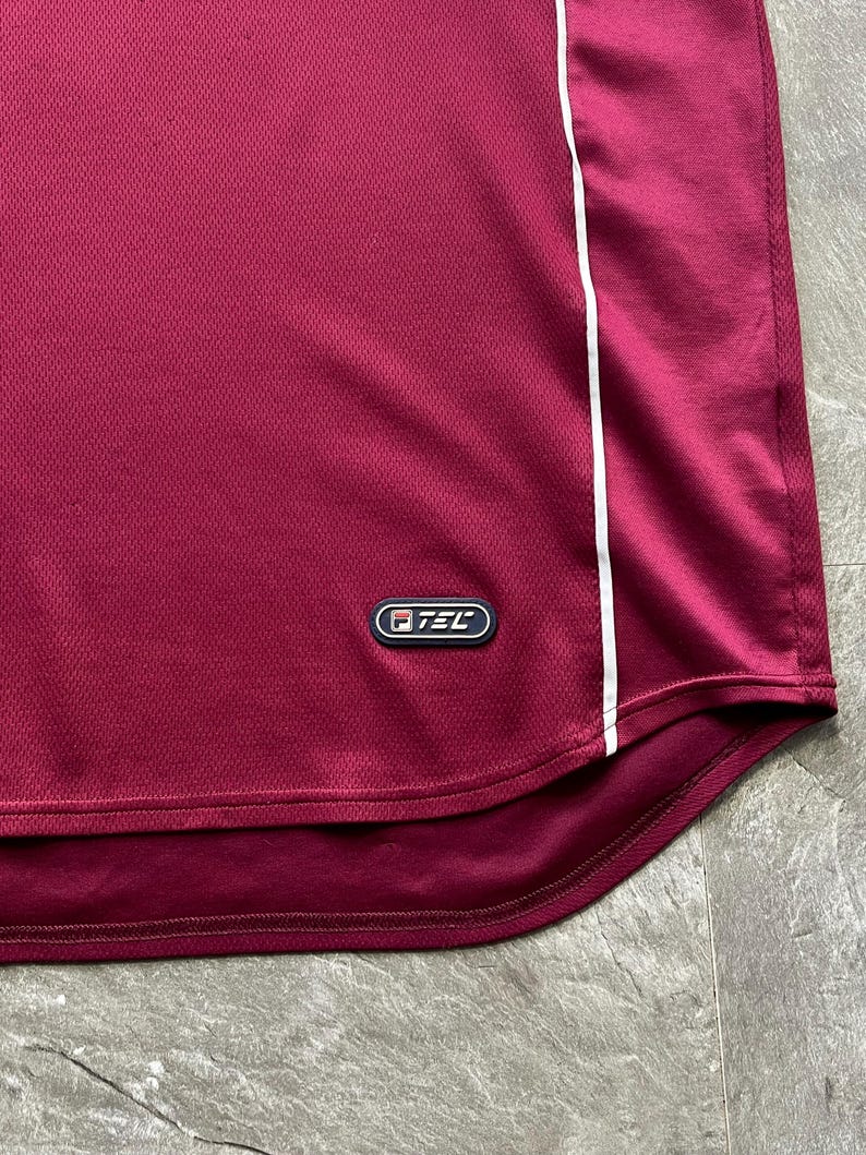 May include: Maroon athletic shirt with a white stripe down the side. The shirt has a mesh texture and a small logo near the bottom. The fabric appears to be a lightweight, breathable material, suitable for sports or casual wear.