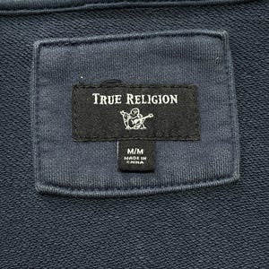 May include: Close-up of a navy blue hoodie label. The label reads "TRUE RELIGION" in white text, with a graphic of a person playing a guitar. Below the logo is a smaller label that reads "M/M MADE IN CHINA".