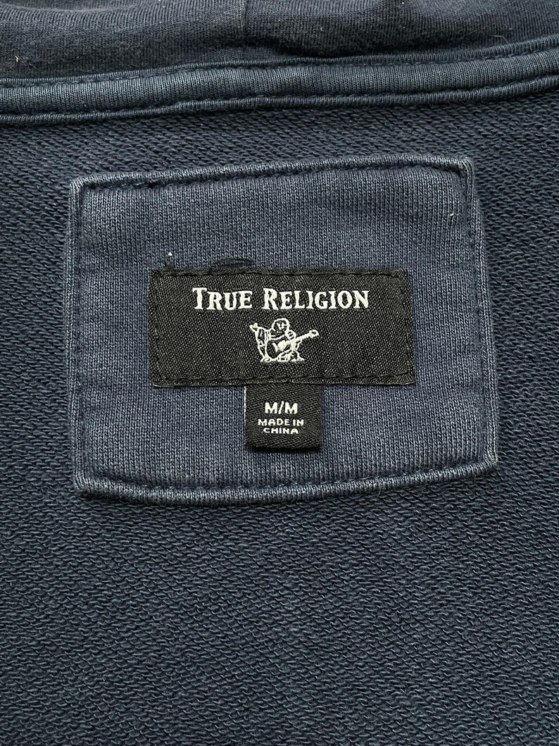 May include: Close-up of a navy blue hoodie label. The label reads "TRUE RELIGION" in white text, with a graphic of a person playing a guitar. Below the logo is a smaller label that reads "M/M MADE IN CHINA".
