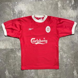 May include: Red short-sleeved jersey with white collar and cuffs. Features the Carlsberg logo in white across the chest, a Reebok logo, and a team crest. The jersey is likely a vintage soccer or football team shirt.