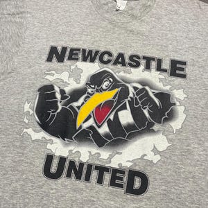 Puede incluir: Camiseta gris con la inscripci&oacute;n "NEWCASTLE UNITED" arriba y abajo de un dibujo de un p&aacute;jaro. El p&aacute;jaro es negro con pico amarillo y detalles rojos, sobre un fondo blanco con forma de nube.