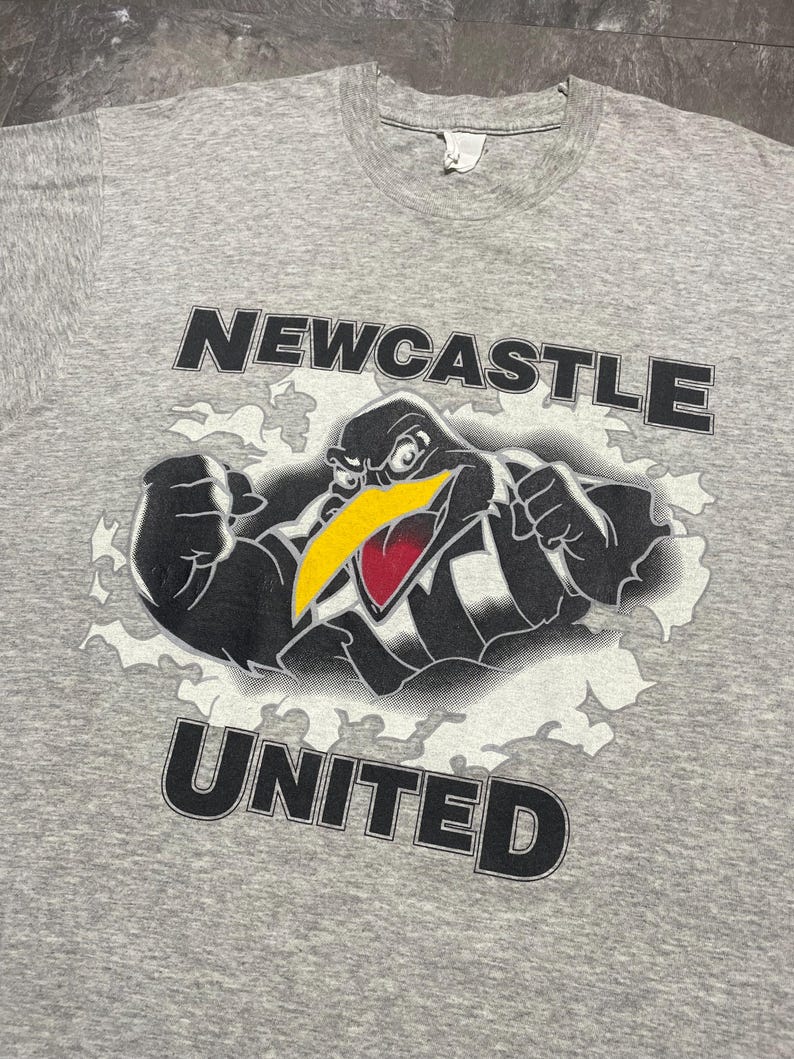 Puede incluir: Camiseta gris con la inscripci&oacute;n "NEWCASTLE UNITED" arriba y abajo de un dibujo de un p&aacute;jaro. El p&aacute;jaro es negro con pico amarillo y detalles rojos, sobre un fondo blanco con forma de nube.
