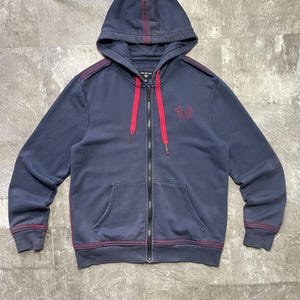 May include: Navy blue zip-up hoodie with a hood and front pockets. The hoodie features red stitching details along the seams and a red drawstring. A small embroidered design is on the left chest.