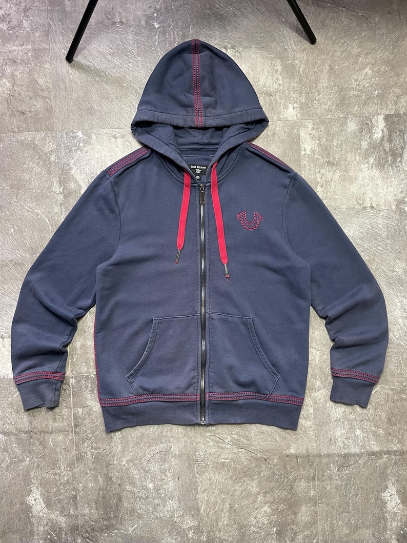 May include: Navy blue zip-up hoodie with a hood and front pockets. The hoodie features red stitching details along the seams and a red drawstring. A small embroidered design is on the left chest.
