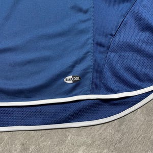 K&ouml;nnte beinhalten: Nahaufnahme eines blauen Sportkleidungsst&uuml;cks mit wei&szlig;em Besatz. Das Kleidungsst&uuml;ck hat einen Mesh-Einsatz und das "CLIMACOOL"-Logo. Der Stoff hat ein strukturiertes Aussehen, und das Gesamtdesign deutet auf Sportbekleidung hin.