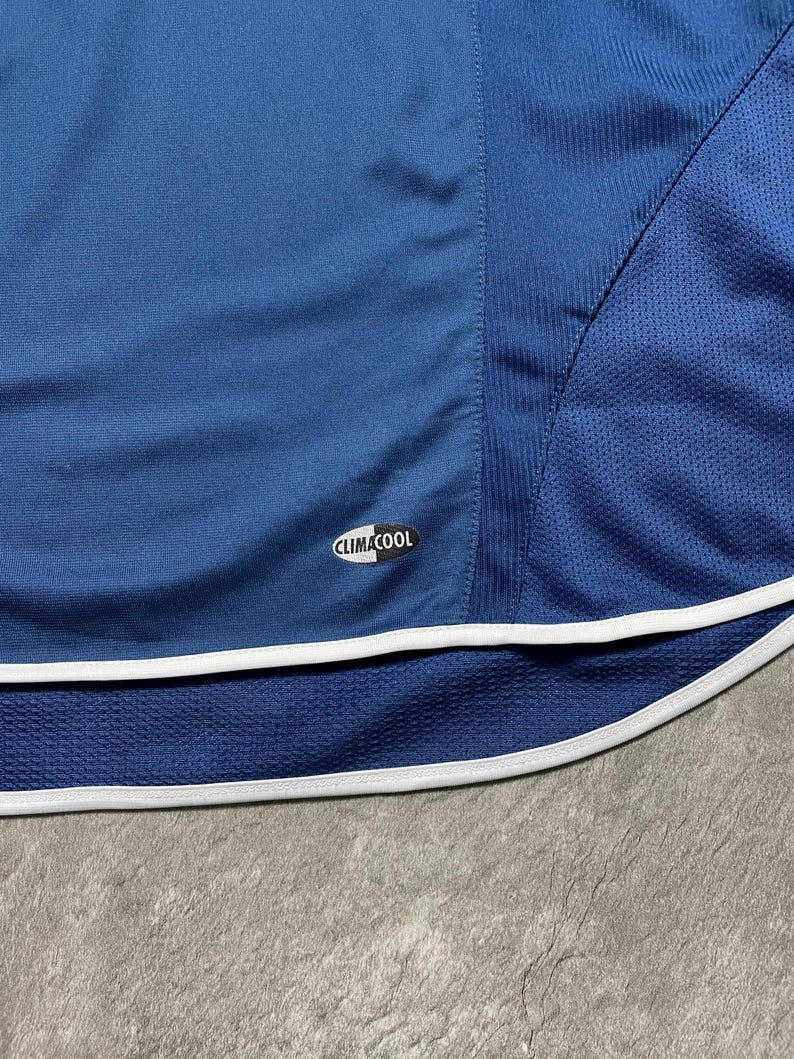 K&ouml;nnte beinhalten: Nahaufnahme eines blauen Sportkleidungsst&uuml;cks mit wei&szlig;em Besatz. Das Kleidungsst&uuml;ck hat einen Mesh-Einsatz und das "CLIMACOOL"-Logo. Der Stoff hat ein strukturiertes Aussehen, und das Gesamtdesign deutet auf Sportbekleidung hin.