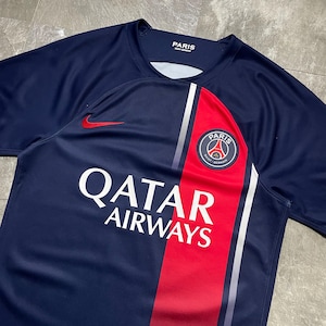 May include: A navy blue short-sleeved jersey featuring a vertical red stripe on the right side with a white border. The shirt displays the Paris Saint-Germain crest, a red Nike swoosh logo, and the white text Qatar Airways on the front chest area.