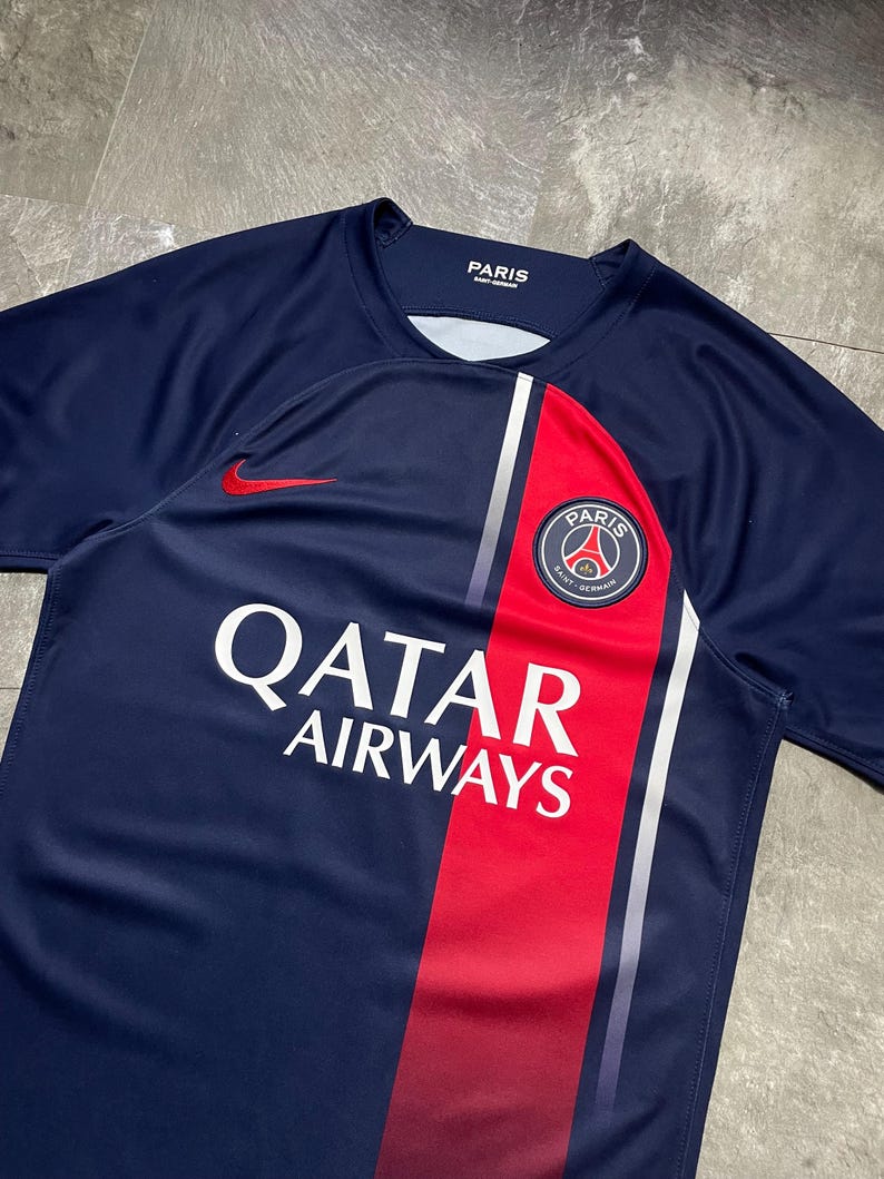 May include: A navy blue short-sleeved jersey featuring a vertical red stripe on the right side with a white border. The shirt displays the Paris Saint-Germain crest, a red Nike swoosh logo, and the white text Qatar Airways on the front chest area.