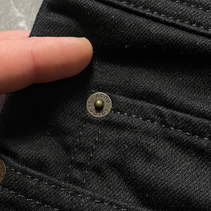 May include: Close-up shot of black denim fabric showing detailed stitching and a brass button. The button is embossed with the text "ACNE STUDIOS". The image focuses on the texture and construction of the garment.