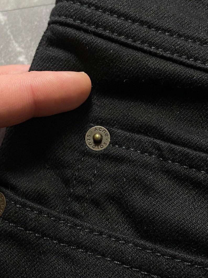 May include: Close-up shot of black denim fabric showing detailed stitching and a brass button. The button is embossed with the text "ACNE STUDIOS". The image focuses on the texture and construction of the garment.