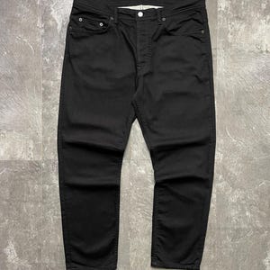 May include: Black denim jeans with a button fly and five pockets. The jeans have a straight-leg cut. The fabric looks to be a medium-weight denim. The jeans are displayed flat on a grey surface.