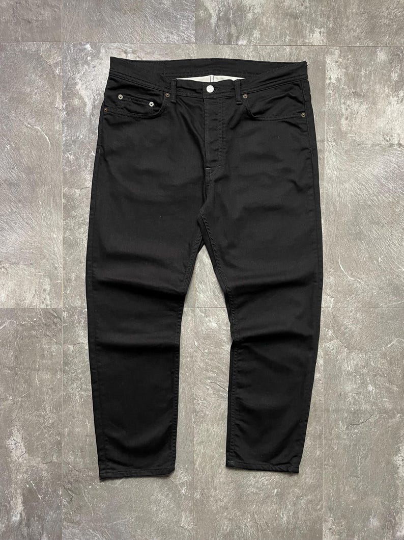 May include: Black denim jeans with a button fly and five pockets. The jeans have a straight-leg cut. The fabric looks to be a medium-weight denim. The jeans are displayed flat on a grey surface.