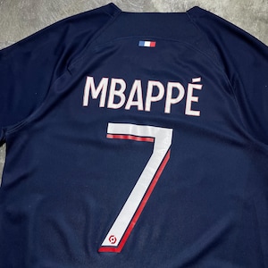 May include: A dark blue sports jersey featuring the name MBAPP&Eacute; printed in white with a thin red outline above the number 7 in white with a red accent. A small French flag emblem is positioned centred above the name on the upper back area.