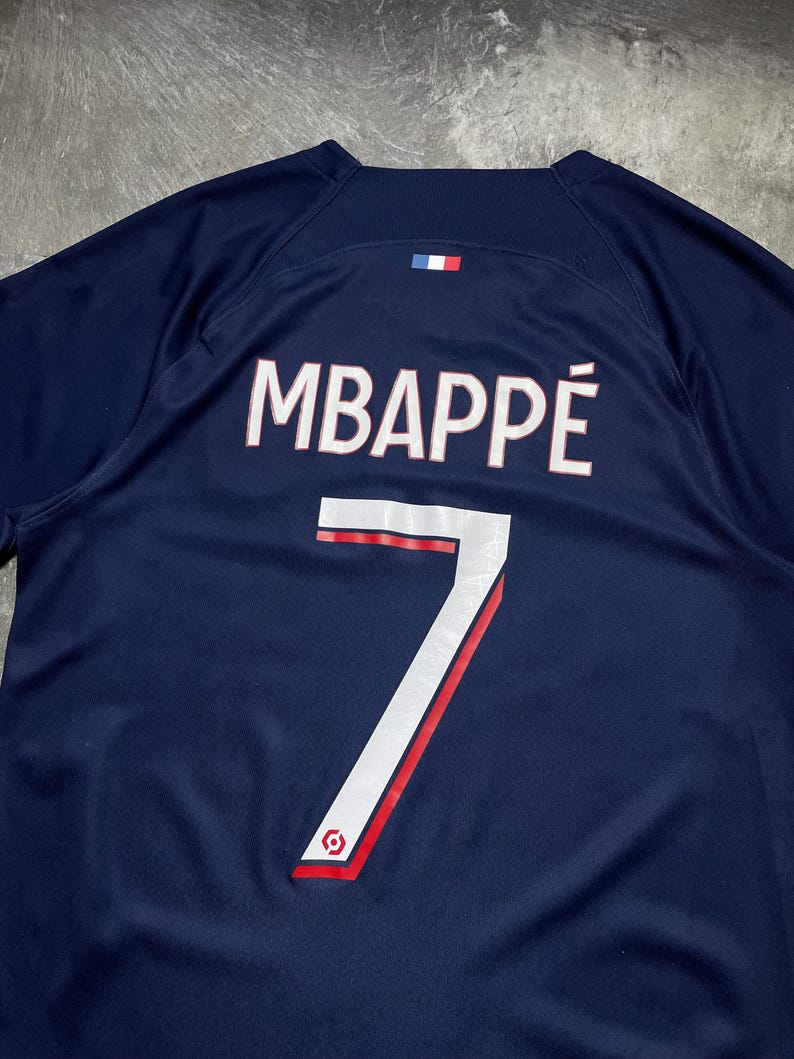 May include: A dark blue sports jersey featuring the name MBAPP&Eacute; printed in white with a thin red outline above the number 7 in white with a red accent. A small French flag emblem is positioned centred above the name on the upper back area.