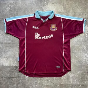 May include: Maroon football shirt with light blue collar and shoulder detailing. Features the text "Dr. Martens" and the Fila logo. The team crest is on the upper right chest. White trim accents the design.