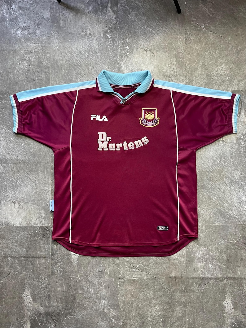May include: Maroon football shirt with light blue collar and shoulder detailing. Features the text "Dr. Martens" and the Fila logo. The team crest is on the upper right chest. White trim accents the design.
