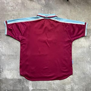 May include: Maroon and light blue short-sleeved jersey. The jersey features a white stripe on the sleeves and collar. The collar is white with maroon and light blue trim. The jersey is made of a shiny material.