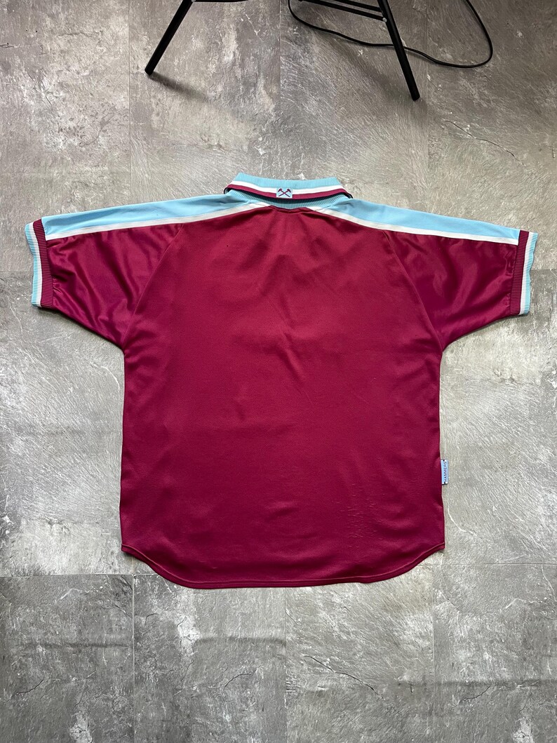 May include: Maroon and light blue short-sleeved jersey. The jersey features a white stripe on the sleeves and collar. The collar is white with maroon and light blue trim. The jersey is made of a shiny material.