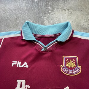 May include: Maroon football shirt with a light blue collar and white trim. Includes the Fila logo, "Dr. Martens" text, and the West Ham United crest. The shirt has a buttoned collar.