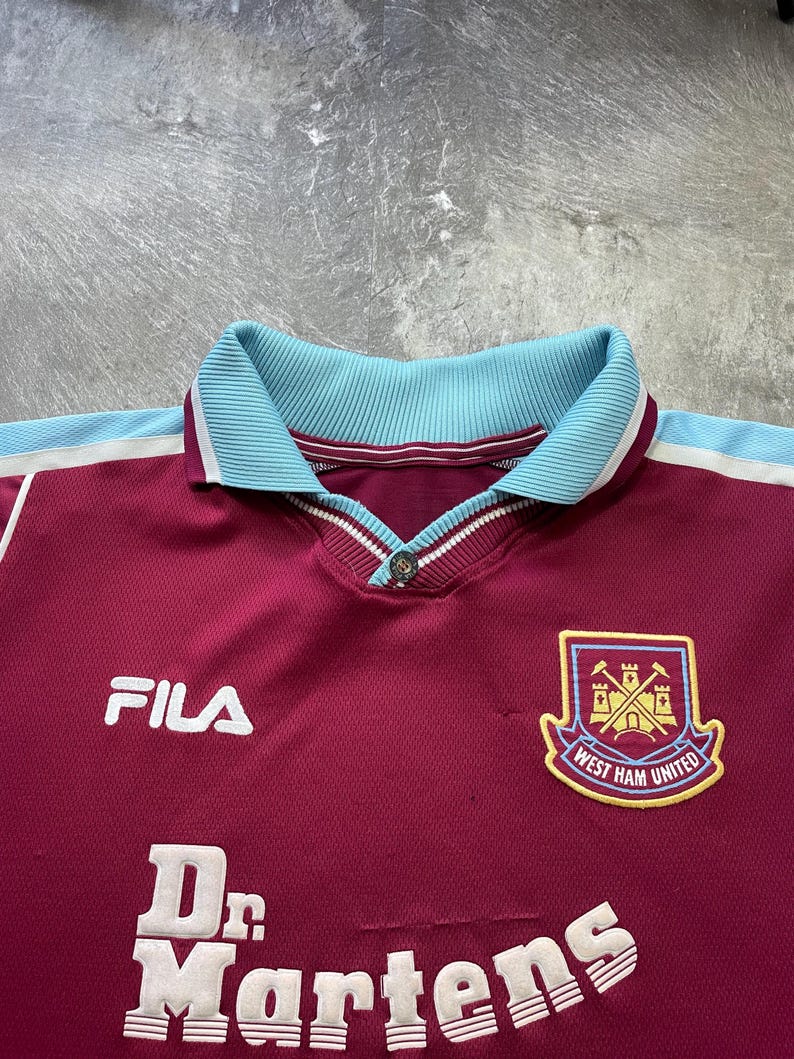 May include: Maroon football shirt with a light blue collar and white trim. Includes the Fila logo, "Dr. Martens" text, and the West Ham United crest. The shirt has a buttoned collar.