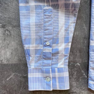 May include: A long-sleeved shirt sleeve featuring a light blue and white plaid pattern. The sleeve includes a buttoned cuff and a placket with a single visible button, showcasing a structured design with clean stitching and a classic woven fabric texture.