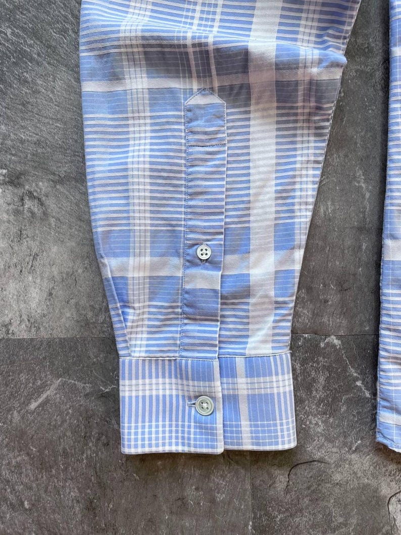 May include: A long-sleeved shirt sleeve featuring a light blue and white plaid pattern. The sleeve includes a buttoned cuff and a placket with a single visible button, showcasing a structured design with clean stitching and a classic woven fabric texture.