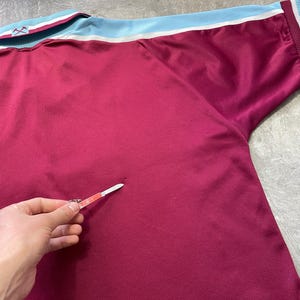 May include: Maroon sports jersey with light blue and white trim on the collar and sleeves. The jersey features a textured fabric and a logo on the collar. A sports-themed garment.