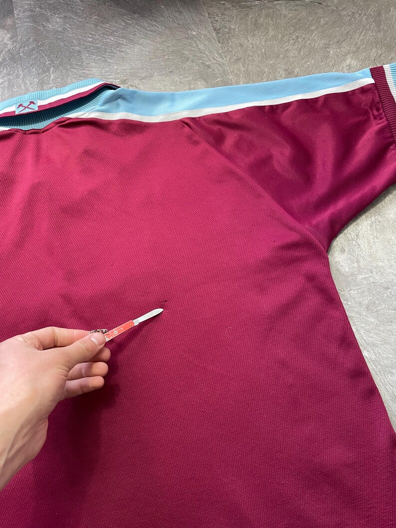 May include: Maroon sports jersey with light blue and white trim on the collar and sleeves. The jersey features a textured fabric and a logo on the collar. A sports-themed garment.