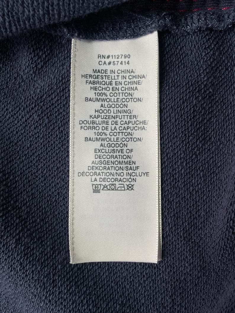 May include: Close-up of a navy blue fabric with a white tag attached. The tag includes text in multiple languages, including "MADE IN CHINA" and fabric composition details. The tag also lists RN and CA numbers.